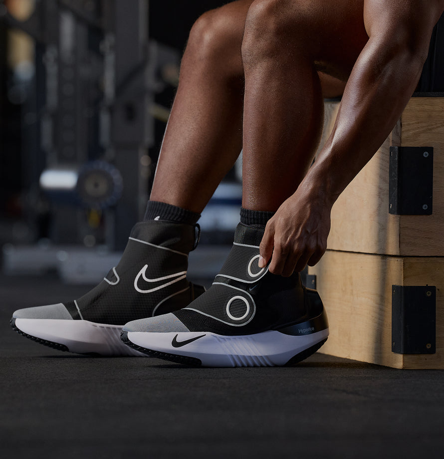 Person wearing Nike x Hyperice Hyperboots sitting on a wooden box in a gym with their feet on the floor.