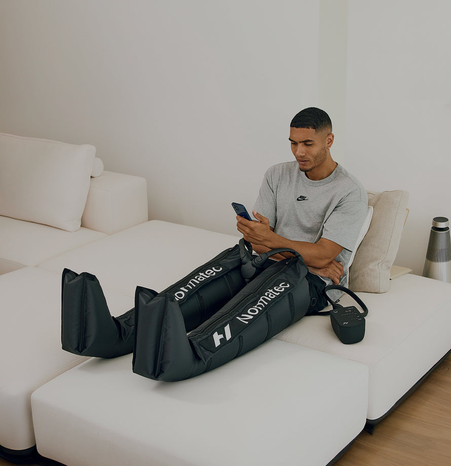 Man sitting on a white couch using a smartphone while he is wearing the Normatec 3 Dymanic air compression leg system