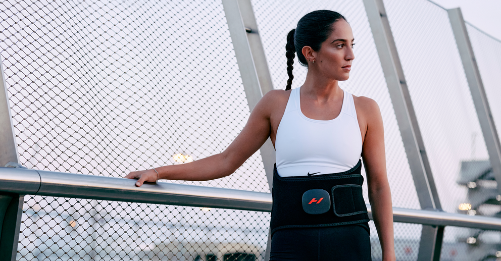 Woman standing on a bridge, wearing a white tank top with black shorts while using a Hyperice Venom 2 Back massager